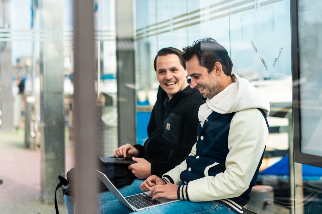 Two people sitting side by side outdoors behind glass, laptops on their laps, sharing a collaborative moment.